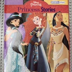 Può includere: Un libro per bambini intitolato "Disney Princess Princess Stories" con illustrazioni delle principesse Disney Jasmine, Merida e Pocahontas. La copertina è rossa e viola con le parole "Princess Stories" in un font grande.