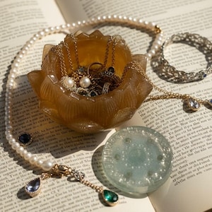 Puede incluir: Un libro abierto con joyas, incluyendo un collar de perlas, cadenas de oro y una pulsera de plata. Un cuenco decorativo color ámbar contiene anillos y collares. Una pieza redonda de vidrio azul claro descansa en la página.