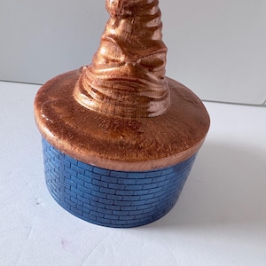 May include: A round container with a blue brick-patterned base and a metallic copper-colored lid shaped like a pointed, wrinkled hat. The lid features deep folds and creases, while the base has a uniform, textured grid of rectangular bricks.