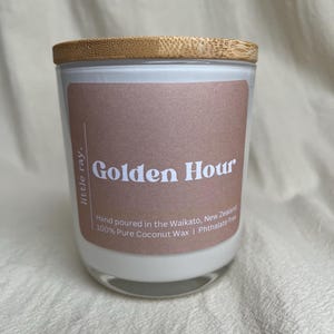 May include: A glass candle jar with a wooden lid. The label reads "Golden Hour" in white text on a brown background. The candle is filled with white wax and the text "Hand poured in the Waikato, New Zealand" is also visible.
