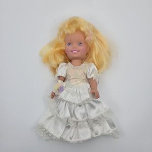 May include: A doll with long blonde hair, blue eyes, and a light skin tone. The doll wears a white satin dress featuring short puff sleeves, a bodice with an iridescent sheen, and a multi-tiered skirt trimmed with white lace and a small floral accent.