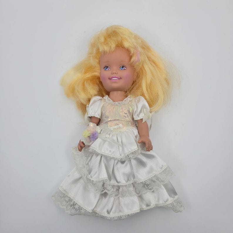 May include: A doll with long blonde hair, blue eyes, and a light skin tone. The doll wears a white satin dress featuring short puff sleeves, a bodice with an iridescent sheen, and a multi-tiered skirt trimmed with white lace and a small floral accent.