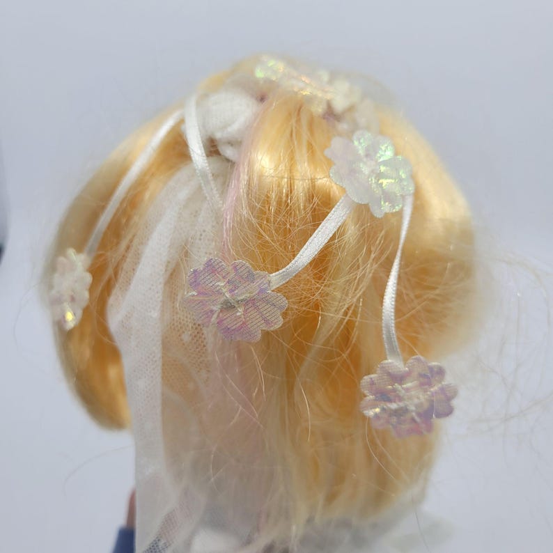 May include: A doll head with blonde hair styled with thin white ribbons and small, iridescent, flower-shaped embellishments. A piece of white, dotted tulle fabric is draped among the hair strands, creating a layered, decorative effect.