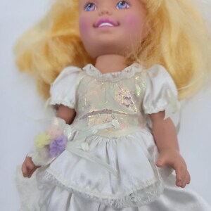 May include: A doll with blonde hair and blue eyes wearing a white dress with a shimmering iridescent bodice, short puffed sleeves, and a lace-trimmed skirt. The doll holds a small bouquet of pastel-coloured pom-poms tied with a white ribbon.