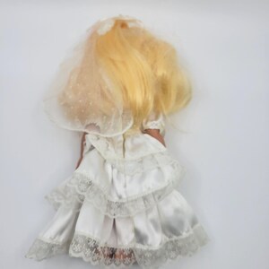 May include: A doll with long blonde hair wearing a white satin wedding dress. The dress features multiple tiered layers with delicate white lace trim along the edges. A sheer, white, polka-dot patterned veil is attached to the back of the hair.