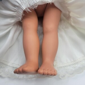 May include: A doll with light tan skin tone legs and feet, wearing a white dress with a gathered waist and a lace-trimmed hem. The fabric of the dress is visible around the doll's legs, showing the texture of the white material and the lace detail.