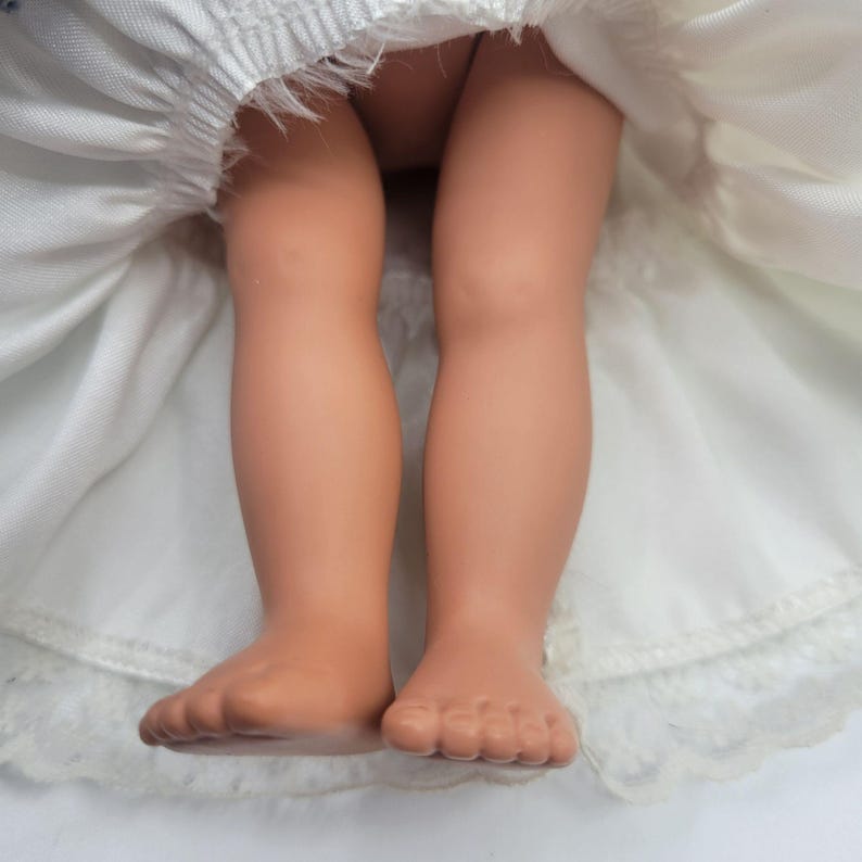 May include: A doll with light tan skin tone legs and feet, wearing a white dress with a gathered waist and a lace-trimmed hem. The fabric of the dress is visible around the doll's legs, showing the texture of the white material and the lace detail.