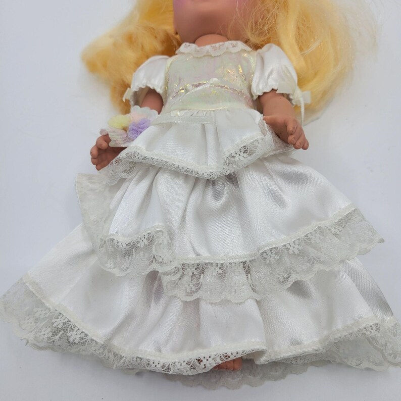 May include: A doll with long blonde hair wearing a white satin dress featuring three tiered ruffled layers trimmed with white lace. The bodice has a shimmering iridescent overlay and short puffed sleeves. A small cluster of pastel pom-poms is held.