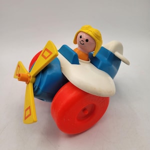 May include: A vintage toy airplane with a yellow propeller, red wheels, and a blue and white body. A small figure with a yellow helmet sits in the cockpit. The toy is designed for children.