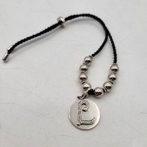May include: Silver-toned necklace with a black cord. The necklace features a circular pendant with the letter "L" encrusted with small, clear crystals. The cord is adjustable with silver-toned beads and a sliding clasp.