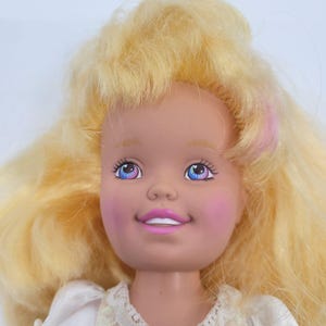 May include: A doll with voluminous blonde hair, light skin, and pink blush on the cheeks. The doll features blue eyes with pink accents, a bright pink smile showing white teeth, and is wearing a white garment with a textured lace collar.