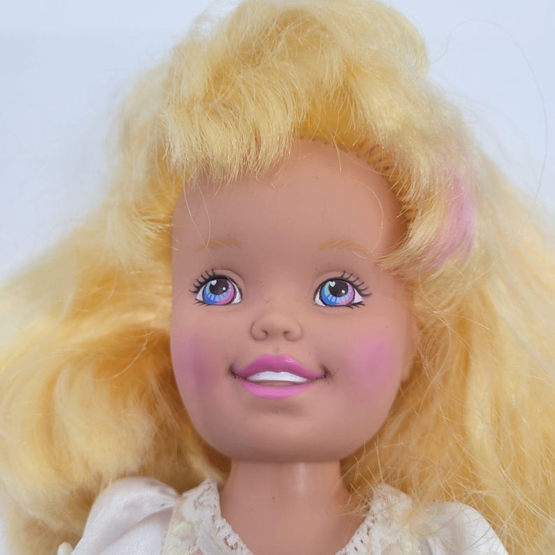 May include: A doll with voluminous blonde hair, light skin, and pink blush on the cheeks. The doll features blue eyes with pink accents, a bright pink smile showing white teeth, and is wearing a white garment with a textured lace collar.
