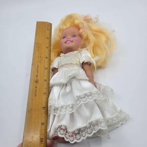 May include: A doll with blonde hair wearing a white dress with tiered ruffles and lace trim, positioned next to a wooden ruler showing a height of approximately 24 cm from the top of the head to the bottom of the dress hemline.