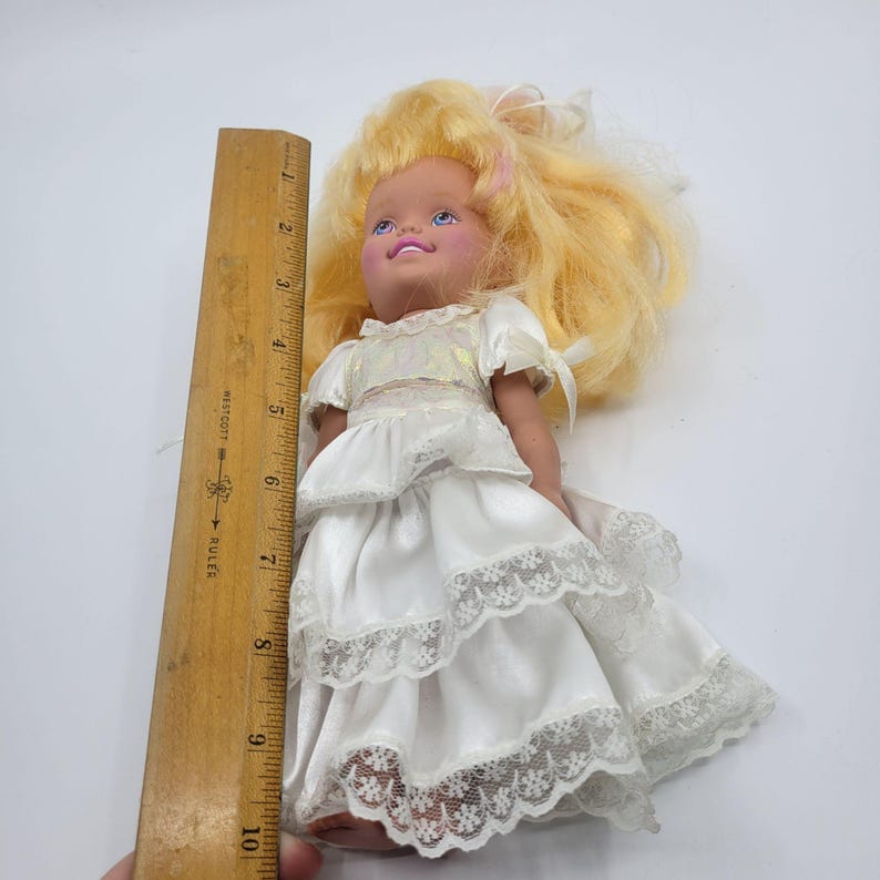 May include: A doll with blonde hair wearing a white dress with tiered ruffles and lace trim, positioned next to a wooden ruler showing a height of approximately 24 cm from the top of the head to the bottom of the dress hemline.