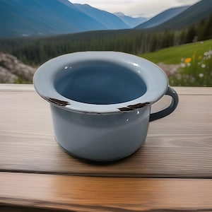 May include: A light blue enamel chamber pot with a handle. The rim has a distressed, chipped appearance. The chamber pot is sitting on a wooden surface, with a scenic mountain backdrop.