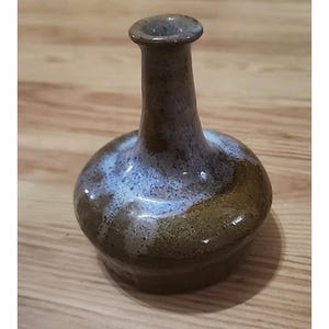 May include: A small ceramic bud vase with a bulbous base and a narrow neck. The lower portion of the vase features a glossy olive-brown glaze, while the upper neck and shoulder area are finished with a speckled, light blue and grey glaze.