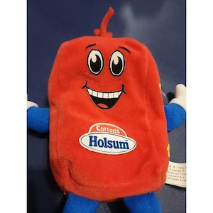 May include: A red plush toy shaped like a loaf of bread with blue arms and legs, white gloves, and a smiling face. The front features a white oval patch with the blue text Cotton's Holsum. A small white tag is attached to the side of the toy.