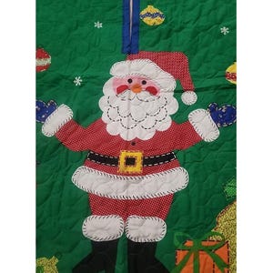May include: A green quilted fabric tree skirt featuring a central applique of Santa Claus wearing a red and white polka dot suit with a black belt and yellow buckle. The background includes embroidered white snowflakes and colorful ornaments.