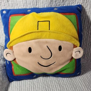 Op de afbeelding: Een vierkant pluche kussen met een stripfiguur met een gele helm, een beige huid en een glimlachend gezicht. Het personage is gecentreerd op een groene vierkante achtergrond met een rode rand, op een blauw veld met zilveren schroefpictogrammen.