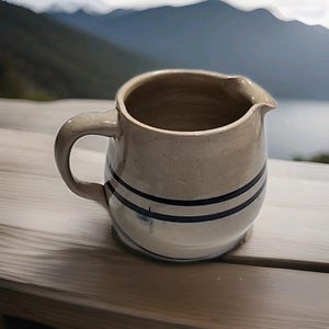 Puede incluir: Una jarra de gres beige con asa y pico vertedor. Dos rayas azul oscuro rodean el cuerpo de la jarra. La jarra está sobre una superficie de madera con un fondo borroso de montaña y lago.