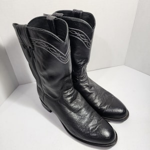 Könnte beinhalten: Ein Paar schwarze Cowboystiefel aus Leder mit strukturierter Oberfläche, spitzer Zehenpartie und einem dekorativen weißen Stickmuster am Schaft. Jeder Stiefel verfügt über zwei Zuglaschen am oberen Rand und einen niedrigen Absatz.