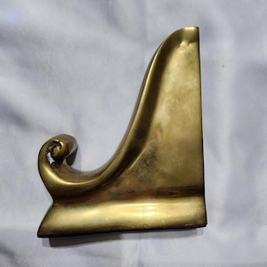 Peut inclure: Un support de présentation en laiton massif avec une finition dorée lisse et polie. L'objet présente une base large et plate ainsi qu'un bras de support incurvé qui s'élève vers le haut et se termine par un détail en volute décorative.