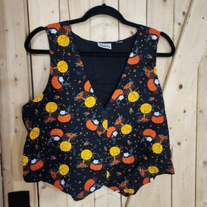 May include: A black sleeveless vest featuring a repeating pattern of orange pumpkins, yellow full moons, and black and white cats. The garment has a V-neckline, a front button closure, and a label inside the collar that reads Basic Editions.