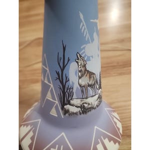 May include: A light blue vase featuring a hand-painted scene of a wolf howling on a rocky outcrop near a bare tree. The base and lower section are decorated with white geometric triangular patterns and linear designs against the blue surface.