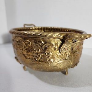 Può includere: Una ciotola rotonda in ottone con un design in rilievo di grappoli d'uva e foglie sull'esterno. La ciotola presenta un bordo con trama a corda, due manici laterali decorativi e poggia su piccoli piedini integrati.