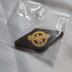 May include: A stack of diamond-shaped black fabric patches enclosed in a clear plastic bag. Each patch features a gold-colored embroidered emblem of an eagle with outstretched wings centered within a circular border, creating a detailed graphic.