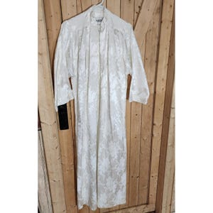 Pode incluir: Um roupão comprido até ao chão com mangas compridas num padrão floral jacquard em creme e branco sujo. A peça apresenta um fecho de correr frontal a todo o comprimento, uma gola alta e uma etiqueta no decote com a inscrição ARIE.