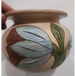 May include: A small, round, beige ceramic pot featuring a carved and painted floral design. The petals are colored in shades of light blue and brown, while a prominent green leaf is depicted on the side. The pot has a wide, flared rim.