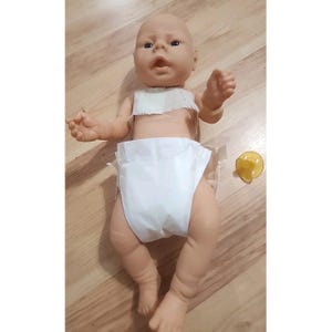 May include: A light-skinned baby doll with molded hair, open eyes, and an open mouth. The doll is wearing a white diaper and a white fabric piece around the chest. A separate yellow plastic pacifier is positioned to the right of the doll.