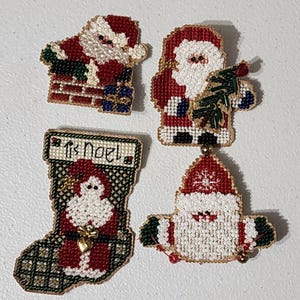 May include: Four handcrafted beaded ornaments featuring Santa Claus designs. One shows Santa in a chimney, another holds a green tree, one is a stocking with the text 'tis Noel', and the last is a Santa head with a red hat and white beard.