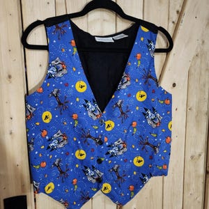 May include: A blue vest featuring a Halloween-themed pattern of orange pumpkins, bare brown trees, yellow full moons with silhouettes of witches on broomsticks, and gray cat faces. The vest has a V-neck, a black interior lining, and three buttons.