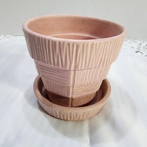 以下が含まれることがあります： 淡いピンク色のセラミック製植木鉢と受け皿のセット。鉢の外側には、縦線と横線を組み合わせたバスケット織り風のテクスチャが施されており、段差のある外観と大きく開いた円形の縁が特徴的です。
