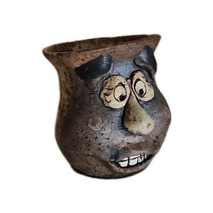 Könnte beinhalten: Ein handgefertigter Keramiktopf mit einem Gesicht auf der Vorderseite. Der Topf ist braun mit schwarzen Akzenten für Augen und Ohren, einer beigen Nase und weißen Zähnen. Der Topf hat ein skurriles, cartoonartiges Design.