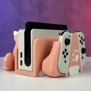 Peut inclure: Un support de console Switch pêche et blanc avec un design sur le thème des chats. Le support maintient la console et les manettes, avec des découpes de pattes et un support en forme de queue. Le support est sur une surface blanche.