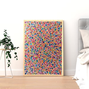 May include: A framed abstract artwork featuring a vibrant pattern of multi-coloured dots. The painting, with hues of blue, pink, yellow, orange, and green, is set against a white wall. The artwork is displayed on a light wooden floor. A small plant is on a side table.