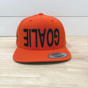 Könnte beinhalten: Orangefarbene Snapback-Kappe mit dem Wort "GOALIE" in großen, schwarzen Buchstaben auf der Vorderseite. Die Kappe hat einen flachen Schirm und einen Druckknopfverschluss auf der Rückseite. Ein kleines Etikett ist auf dem Schirm sichtbar.