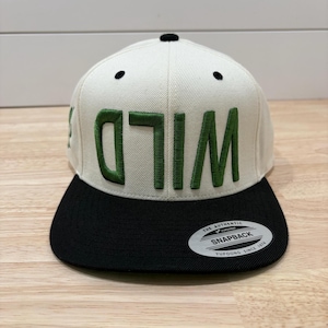 May include: A cream and black snapback cap with the word "WILD" embroidered in green. The cap has a flat peak and a black top. The cap has a "Snapback" tag.