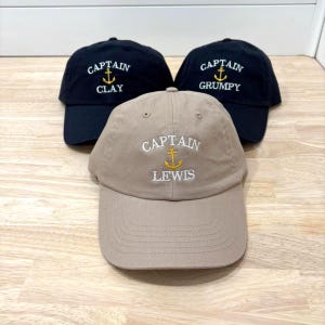 Könnte beinhalten: Drei Baseballkappen mit gesticktem Text und Ankerdesign. Eine Kappe ist beige mit "CAPTAIN LEWIS" in Weiß und Gold. Die anderen beiden sind schwarz, mit "CAPTAIN CLAY" und "CAPTAIN GRUMPY" in Weiß und Gold.