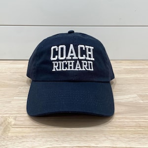 Könnte beinhalten: Marineblaue Baseballkappe mit dem weißen, gestickten Text "COACH RICHARD". Die Kappe hat einen gebogenen Schirm und einen kleinen Knopf oben. Ein klassisches und stilvolles Accessoire.