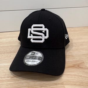 Op de afbeelding: Een zwarte honkbalpet met een wit geborduurd logo op de voorkant bestaande uit de letters S en D in elkaar verweven. De pet heeft een ronde sticker op de klep met de tekst 39THIRTY en LARGE-XLARGE, en een New Era logo aan de zijkant.