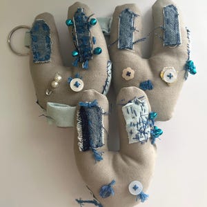 May include: Handmade fabric keychains in the shape of hands. The keychains are beige with denim patches, blue accents, buttons, and small bells. One keychain has a silver keyring attached and a pearl pin.