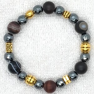 May include: A beaded bracelet featuring a mix of dark gray, gold, and brown beads. The bracelet includes round, patterned, and faceted beads, along with gold-colored spacers. The beads are strung together to form a complete circle.