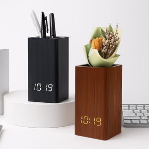 May include: Two wooden desk tidies. One is black and holds pens, the other is brown and holds a small bouquet. Both have a digital clock display showing 10:19.