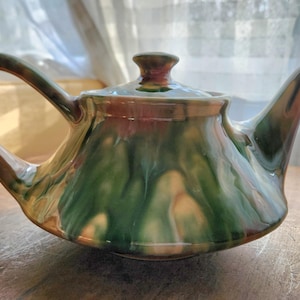 May include: A ceramic teapot with a lid, handle, and spout. The teapot is glazed with a blend of green, brown, and cream colours. The handle curves gracefully, and the spout is pointed. The teapot sits on a wooden surface.