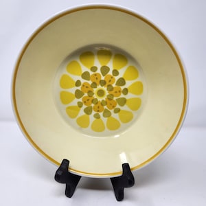 Pode incluir: Um prato redondo de cerâmica creme com um desenho floral amarelo e castanho no centro. O prato tem uma borda amarela e é exibido em um suporte preto. Artigo decorativo.
