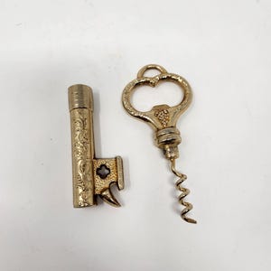 May include: A gold-coloured metal bottle opener shaped like a key with intricate floral engravings and a small dark inset, alongside a matching gold-coloured corkscrew featuring a decorative handle with a grape cluster motif and a spiral metal worm.